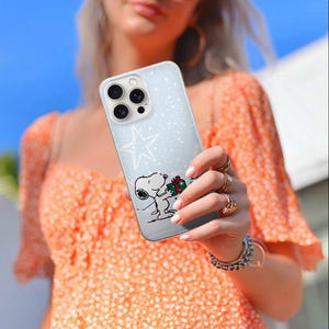 May include: A clear phone case featuring a cartoon Snoopy character holding a gift. The case has a light blue background with a white star and small white dots. The phone case is held by a person wearing rings and bracelets.