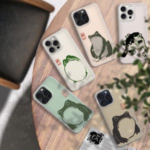 May include: A collection of phone cases with Japanese-inspired frog illustrations in various colors. The cases feature different frog designs in black, green, and white, with a minimalist aesthetic. Some cases have a light green or beige background.