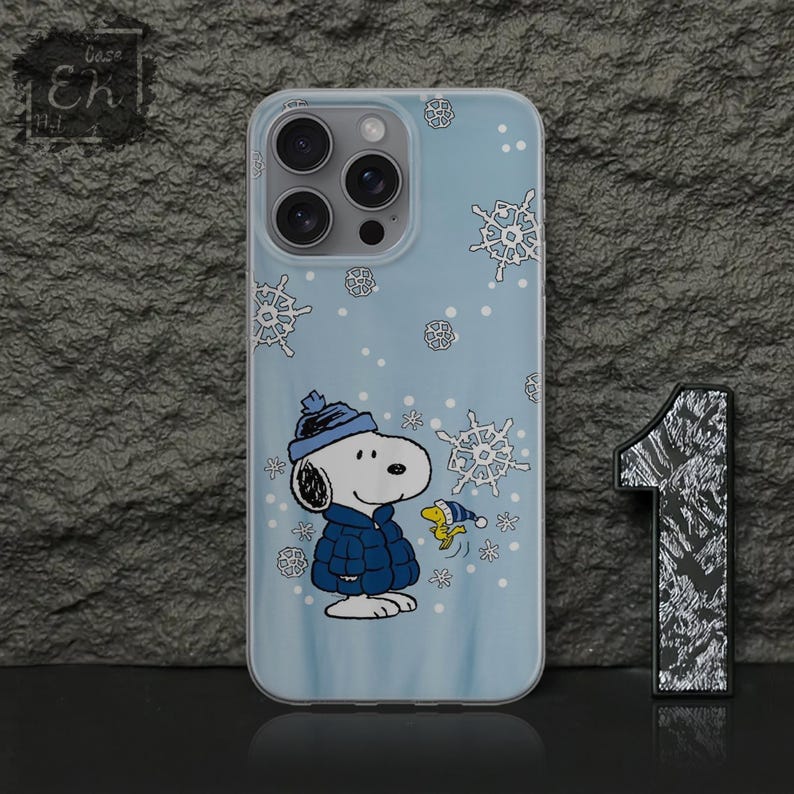 May include: A light blue phone case featuring a cartoon Snoopy character in a winter outfit, surrounded by snowflakes. The case has a clear border and a glossy finish. The Snoopy design includes a blue hat and coat. The case is designed for a smartphone.