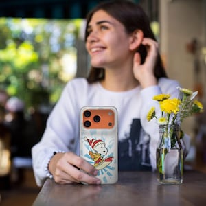 May include: A clear phone case featuring a cartoon Snoopy character wearing a Santa hat and surfing on a surfboard. The design includes a blue and white burst pattern in the background. The phone case has an orange camera section.