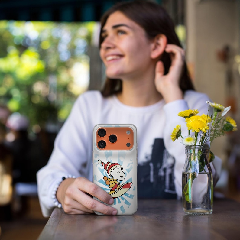 May include: A clear phone case featuring a cartoon Snoopy character wearing a Santa hat and surfing on a surfboard. The design includes a blue and white burst pattern in the background. The phone case has an orange camera section.