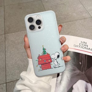 May include: A light blue iPhone case featuring a cartoon Snoopy character with a red doghouse and a Christmas tree. The case has a light blue background with white dots and a clear camera lens area.