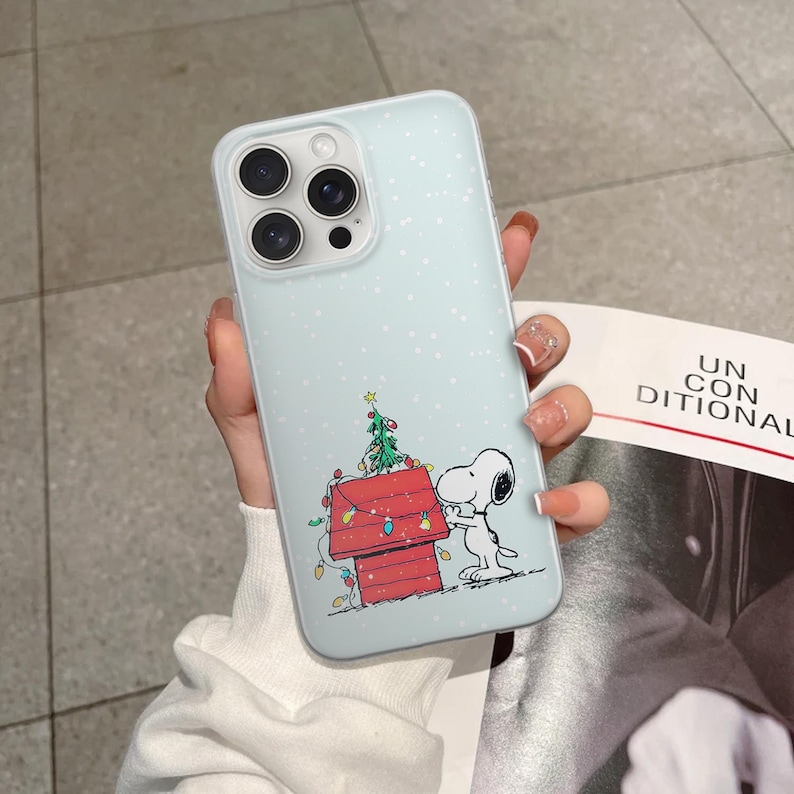 May include: A light blue iPhone case featuring a cartoon Snoopy character with a red doghouse and a Christmas tree. The case has a light blue background with white dots and a clear camera lens area.