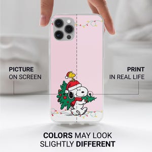 May include: A pink phone case featuring a cartoon Snoopy character wearing a Santa hat and scarf, carrying a Christmas tree. The case has a clear border and a design of Christmas lights. The text at the bottom reads "COLORS MAY LOOK SLIGHTLY DIFFERENT."