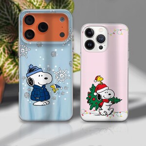May include: Two phone cases featuring Snoopy from Peanuts. One case is light blue with snowflakes and Snoopy in a winter coat. The other is pink with Snoopy carrying a Christmas tree, with a string of lights.