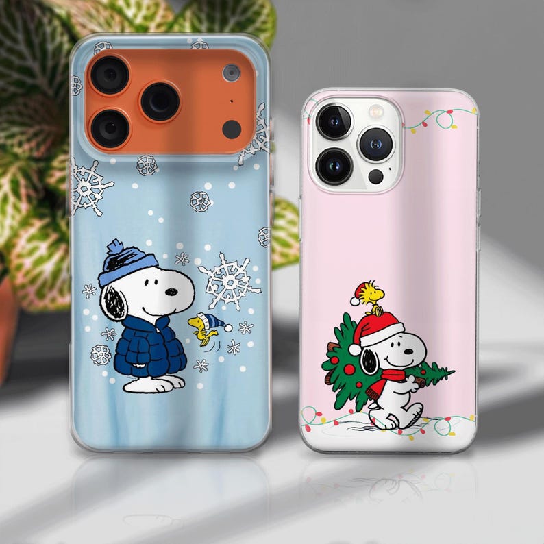 May include: Two phone cases featuring Snoopy from Peanuts. One case is light blue with snowflakes and Snoopy in a winter coat. The other is pink with Snoopy carrying a Christmas tree, with a string of lights.