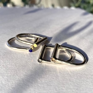 May include: Three silver rings with different coloured stones. The rings are stacked on top of each other. The top ring has a yellow stone, the middle ring has a blue stone, and the bottom ring has a brown stone.