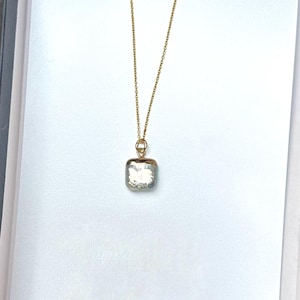Peut inclure: Un collier en chaîne dorée avec un pendentif de forme carrée. Le pendentif est fait d'un matériau gris iridescent. Le collier est présenté dans une boîte blanche avec le texte "SMALL VICTORY JEWELRY" imprimé sur le dessus.