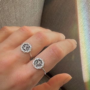 Può includere: Due anelli d'argento con un design geometrico che presenta un triangolo e un simbolo al suo interno. Gli anelli sono al dito di una persona.