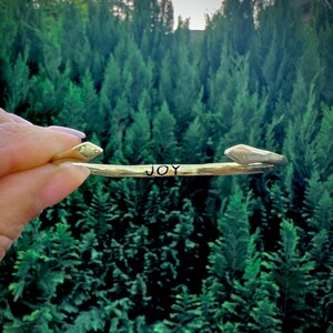 May include: A gold-toned metal bracelet with the word "JOY" engraved on it. The bracelet is shown against a blurred background of green trees.