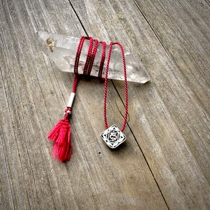 May include: A red cord necklace with a silver pendant featuring a geometric design. The pendant is a square with a circle in the centre and the number "5" inside the circle. The necklace is on a wooden surface with a clear quartz crystal.