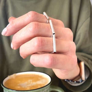 May include: A silver ring with multiple connected bands, worn on a finger. The ring is made of thin, straight pieces of metal that are connected to each other. The ring is worn on the middle finger of a hand.