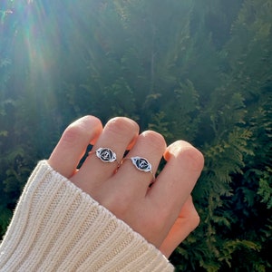 Op de afbeelding: Twee zilveren ringen met een zwart derde oog symbool op elk. De ringen worden gedragen aan de vingers van een hand.