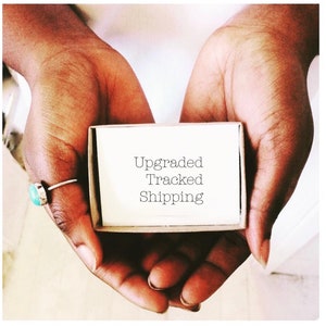 Könnte beinhalten: Ein kleiner, offener Pappkarton mit den Worten "Upgraded Tracked Shipping" auf einem weißen Etikett. Die Schachtel wird in geformten Händen gehalten, mit einem Silberring mit einem Türkisstein an einem Finger.