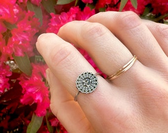 May include: A silver ring with an intricate design featuring the Om symbol in the center. The ring is worn on a finger against a background of pink flowers.