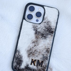 May include: A black phone case with a cowhide design. The case features a black border and a natural white, brown, and black pattern. The initials "KM" are visible on the lower part of the case.