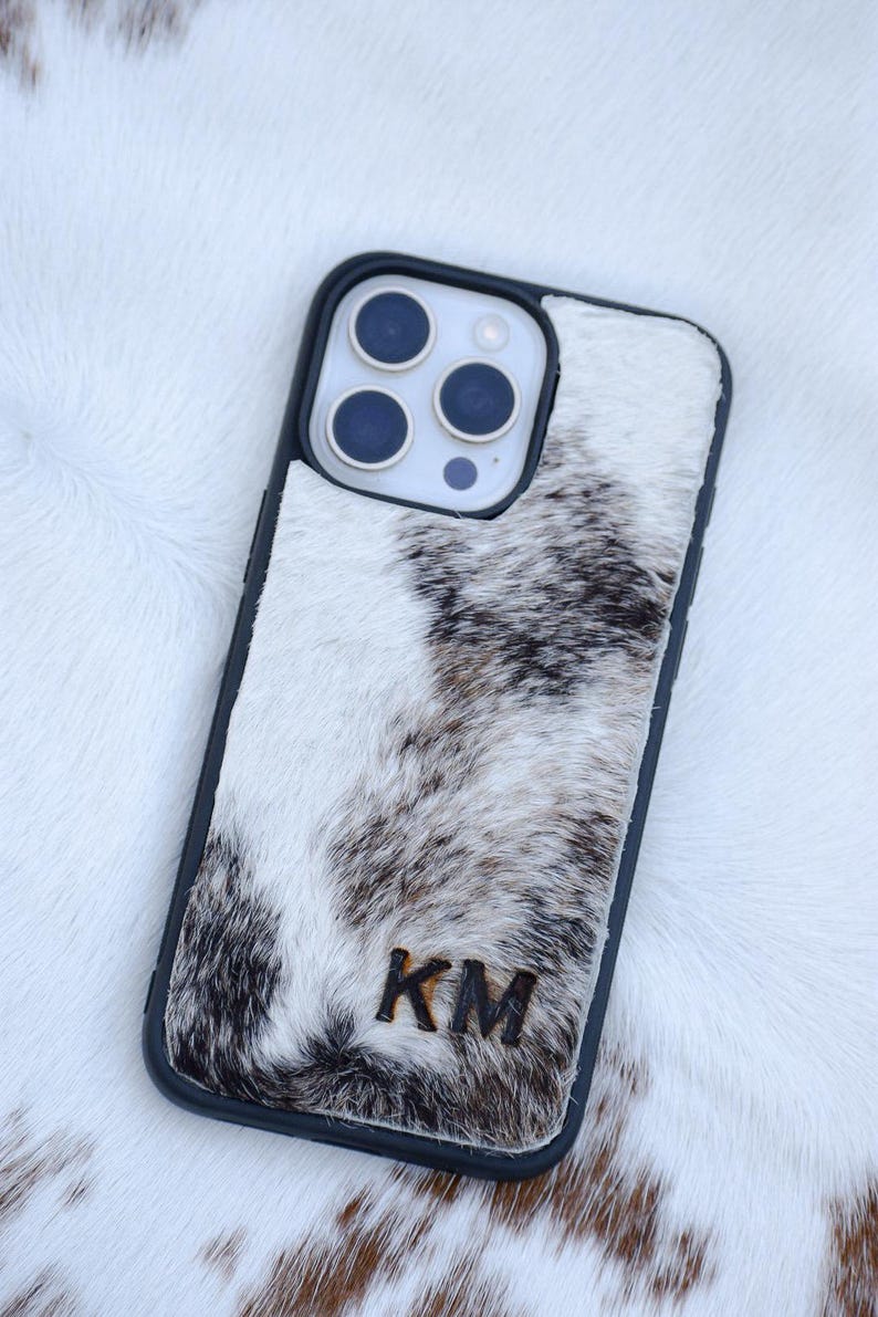 May include: A black phone case with a cowhide design. The case features a black border and a natural white, brown, and black pattern. The initials "KM" are visible on the lower part of the case.
