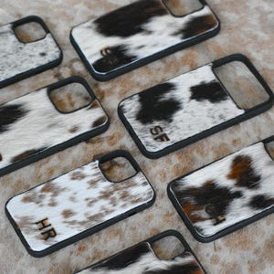 May include: A collection of phone cases with a cowhide pattern. The cases feature a black border and a natural hide design in shades of white, brown, and black. Some cases have initials engraved on them.