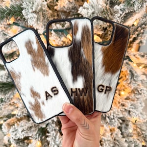 May include: Three phone cases with a cowhide pattern in shades of brown, white, and black. Each case has a black border and initials carved into the hide. The initials are "AS", "HW", and "GP".