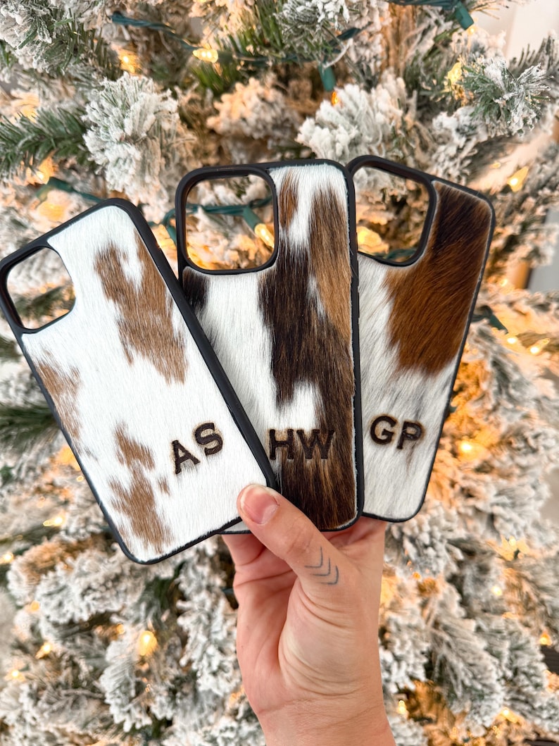 May include: Three phone cases with a cowhide pattern in shades of brown, white, and black. Each case has a black border and initials carved into the hide. The initials are "AS", "HW", and "GP".