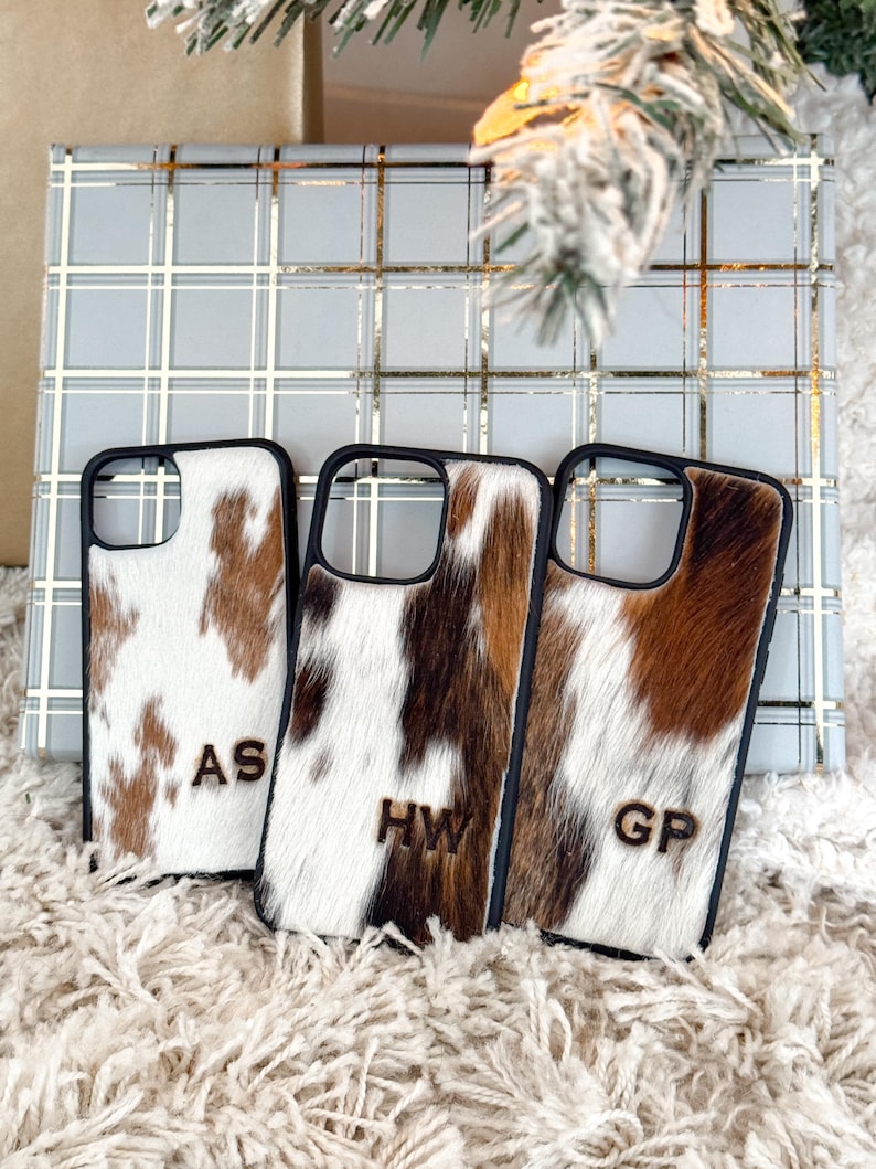 May include: Three personalized phone cases with cowhide patterns. Each case has a black border and features a unique combination of brown and white fur. The cases are monogrammed with the initials "AS", "HW", and "GP" in a wooden font.