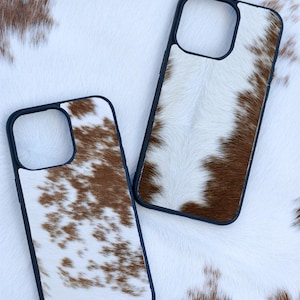 Pu&ograve; includere: Due custodie per telefono con un design in pelle di mucca. Le custodie hanno un bordo nero e un motivo maculato bianco e marrone. Le custodie sono esposte su uno sfondo di pelle di mucca bianca e marrone.