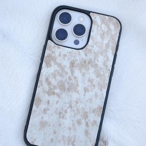 Pu&ograve; includere: Custodia per telefono nera con motivo a pelle di mucca nei toni del bianco e beige. La custodia ha un bordo nero e ritagli per la fotocamera e i pulsanti del telefono. Appoggiata su una superficie bianca simile alla pelliccia.
