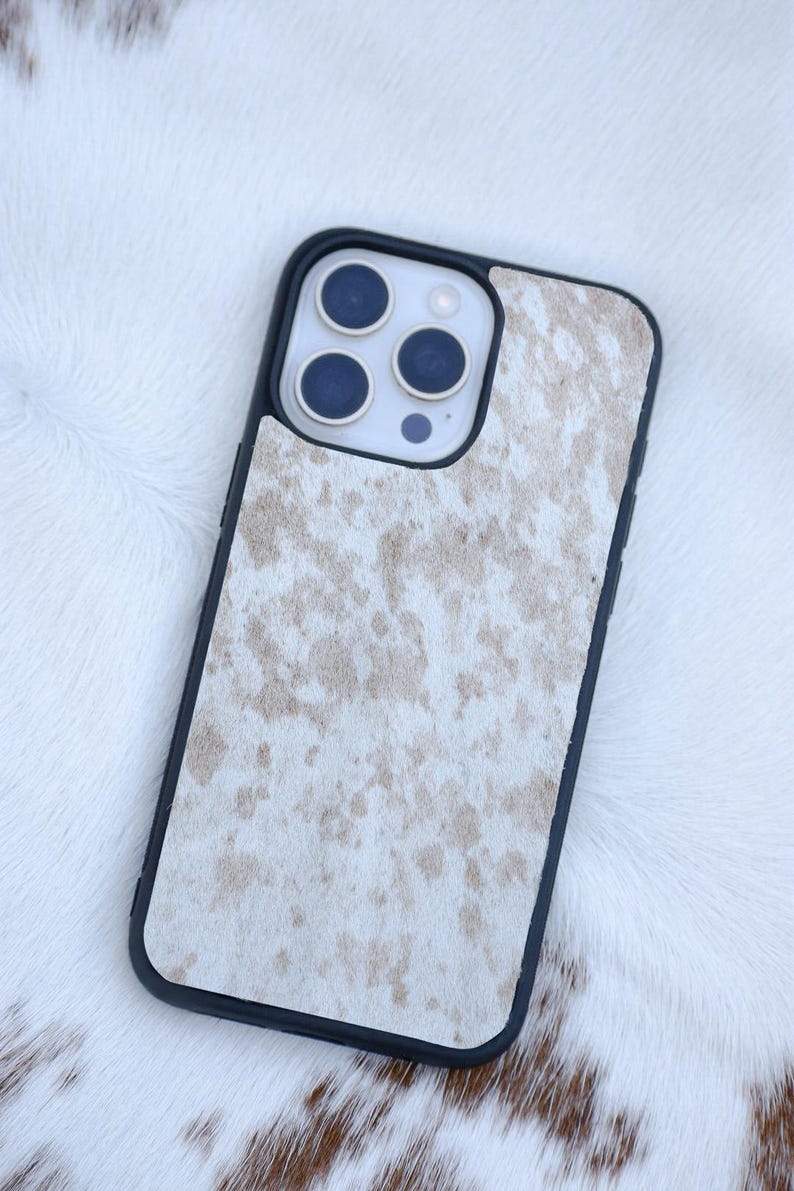 Pu&ograve; includere: Custodia per telefono nera con motivo a pelle di mucca nei toni del bianco e beige. La custodia ha un bordo nero e ritagli per la fotocamera e i pulsanti del telefono. Appoggiata su una superficie bianca simile alla pelliccia.