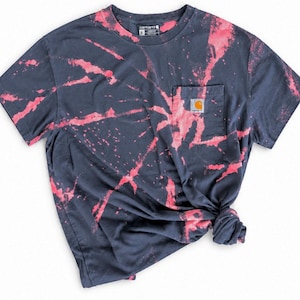 以下が含まれることがあります： ダークチャコールグレーの半袖Tシャツで、ピンクのタイダイ柄が施されています。胸ポケットにはCarharttのロゴパッチがあり、クルーネック仕様です。右下の生地がまとめられ、結び目のような外観になっています。