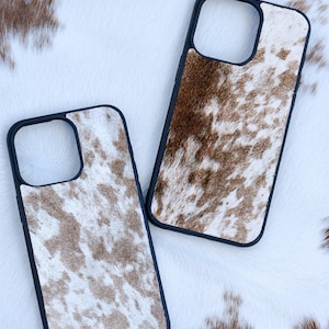 Pu&ograve; includere: Due custodie per telefoni con motivo a pelle di mucca. Le custodie hanno un bordo nero e un design maculato marrone e bianco. Le custodie sono su una superficie di pelle di mucca bianca e marrone.