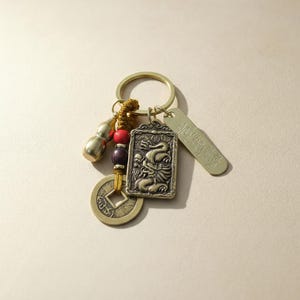 May include: A brass keychain with a rectangular dragon pendant, a round coin, a small gourd, and a tag with text. The keychain also includes red and brown beads and a gold-coloured ring.