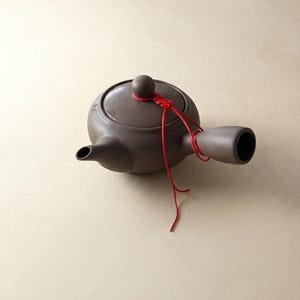 May include: A dark brown ceramic teapot with a side handle and a matching lid. A thin red cord is tied around the lid knob and the handle, featuring a decorative knot. The teapot has a short, tapered spout and a smooth, matte finish throughout.