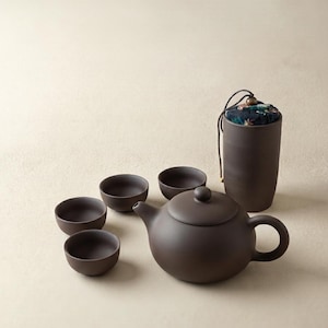 Puede incluir: Un juego de té de cerámica mate en color marrón oscuro que incluye una tetera redonda con tapa, tres tazas pequeñas a juego y un recipiente cilíndrico alto con una tapa de tela estampada asegurada por un cordón marrón y una cuenta.