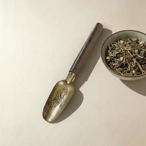 Puede incluir: Una cuchara de té con mango de madera y una cuchara de latón con un diseño grabado. También se ve un pequeño cuenco de cerámica lleno de té de hojas sueltas. La cuchara de té está orientada hacia el cuenco, lo que sugiere su uso para servir té.