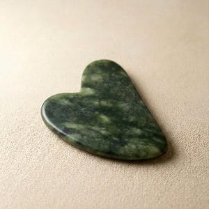 Puede incluir: Una herramienta gua sha de jade verde en forma de corazón. La herramienta tiene una superficie lisa y pulida con variaciones naturales de color. El jade es de un verde oscuro con vetas verdes más claras.