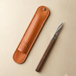 Pode incluir: Uma faca de precisão com cabo de madeira castanho-escuro e lâmina de metal afiada, acompanhada por uma bainha de couro bege com um botão de pressão de latão e um emblema estampado de um símbolo de tridente na parte frontal da capa.
