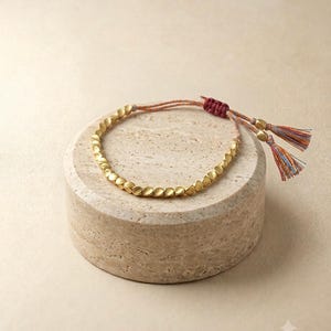 Könnte beinhalten: Ein Armband mit einer Reihe kleiner, mattgoldener Perlen in kreisförmiger Anordnung. Das Armband wird mit einem dunkelroten, verstellbaren Knotenschnurverschluss gesichert und endet in zwei hängenden Quasten aus bunten Fäden und Goldperlen.