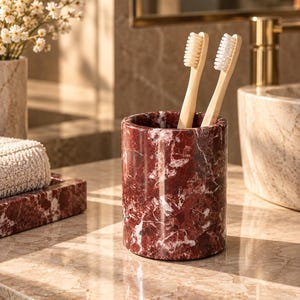 May include: A cylindrical toothbrush holder made of polished dark red marble with white veining. Two bamboo toothbrushes with light-colored bristles are placed upright inside the holder, which features a smooth, glossy finish and a solid base.