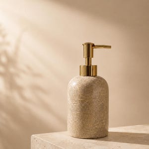 May include: A cylindrical soap dispenser with a textured, speckled beige stone-like body and a polished gold-tone metal pump mechanism. The dispenser features a smooth, rounded top and a flat base, providing a functional and clean aesthetic for any space.