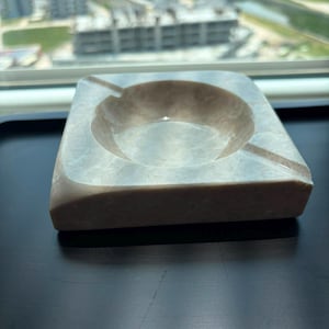 May include: A square, beige marble ashtray with a circular indentation for ashes and a groove for a cigarette. The ashtray is made of polished stone and sits on a dark surface. A smoking accessory.