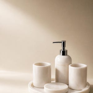 Op de afbeelding: Een badkameraccessoireset gemaakt van gebroken witte steen met een gepolijste afwerking. De set bevat een cilindrische zeepdispenser met een zilverkleurige metalen pomp, twee bijpassende cilindrische bekers en een rond zeepbakje op een dienblad.