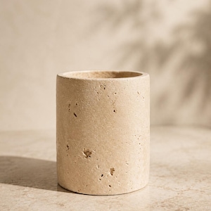 Peut inclure: Un récipient cylindrique fabriqué en pierre de travertin beige, présentant une surface poreuse et texturée avec des piqûres et des variations naturelles sur tout l'extérieur. Le vase possède une base plate et un sommet ouvert à bord épais.