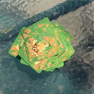 Puede incluir: Una pila de cartas ilustradas verdes y naranjas con una persona sentada en posición de loto rodeada de flores. Las cartas tienen un contorno blanco y el texto "88" en el frente.