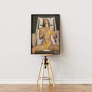 Puede incluir: Una impresión enmarcada de una mujer en bikini amarillo, reclinada en una silla de playa de madera. La obra de arte se exhibe sobre un taburete de madera con base de metal negro, contra una pared blanca y un suelo de madera.