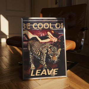 May include: Framed art print featuring a leopard walking towards the viewer, with a woman reclining on a red sofa in the background. The text "BE COOL OR LEAVE" is displayed in gold lettering. The artwork is in a silver frame.