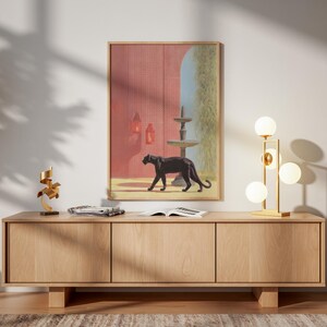 Peut inclure: Une impression encadrée représente une panthère noire devant une fontaine et un mur rouge. Un meuble en bois à trois portes supporte une lampe dorée avec des lumières sphériques. Une sculpture dorée et une chaise sont également visibles.