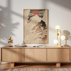 May include: A framed artwork depicting a figure with wings, set against a backdrop of clouds and a red sun. The artwork is displayed above a light wood cabinet with three doors. A gold lamp with spherical bulbs and a small gold sculpture are on the cabinet.