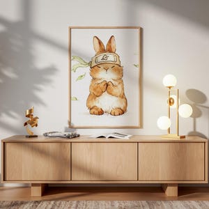 May include: A framed watercolor print of a brown bunny wearing a headband with a symbol. The bunny is in a prayerful pose. The print hangs above a light wood cabinet with a gold lamp and a modern sculpture.