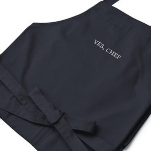 May include: A dark blue apron with adjustable straps and a white embroidered message that says "YES, CHEF".