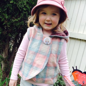 Op de afbeelding: Een jong meisje draagt een roze en blauwe geruite vest met een bruine capuchon. Het vest heeft een knoop met een cartoonmeisje erop. Ze draagt ook een roze hoed en een lang mouwig roze shirt.